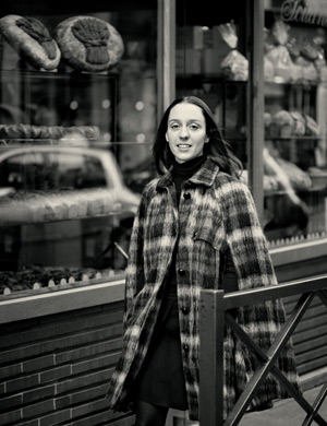 Pictured outside her Paris bakery, Apollonia is wearing a check tweed wool cape by PAUL SMITH, a black cashmere and silk turtleneck sweater by YVES SAINT LAURENT, and a navy and black wool and lace skirt by VANESSA BRUNO with black tights by FALKE.